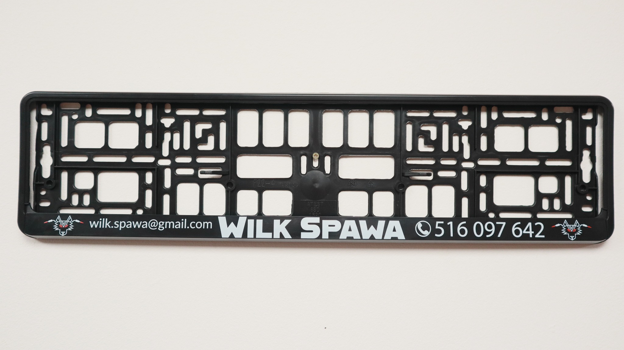 Czarna ramka tablicy rejestracyjnej z nadrukowanymi danymi kontaktowymi firmy Wilk Spawa: wilk.spawa@gmail.com, numer telefonu 516 097 642 oraz grafika wilka po bokach, prezentowana na jasnym tle.