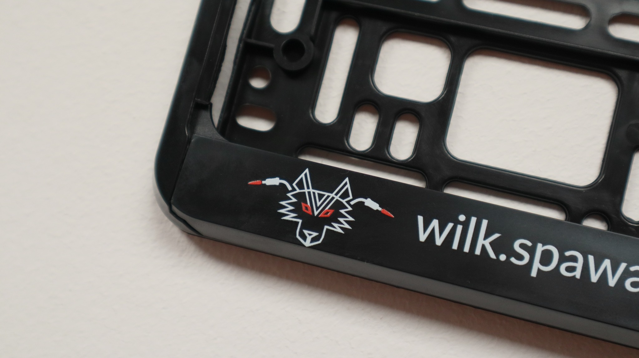 Czarna ramka tablicy rejestracyjnej z białym logo wilka z czerwonymi oczami i napisem 'wilk.spawa'