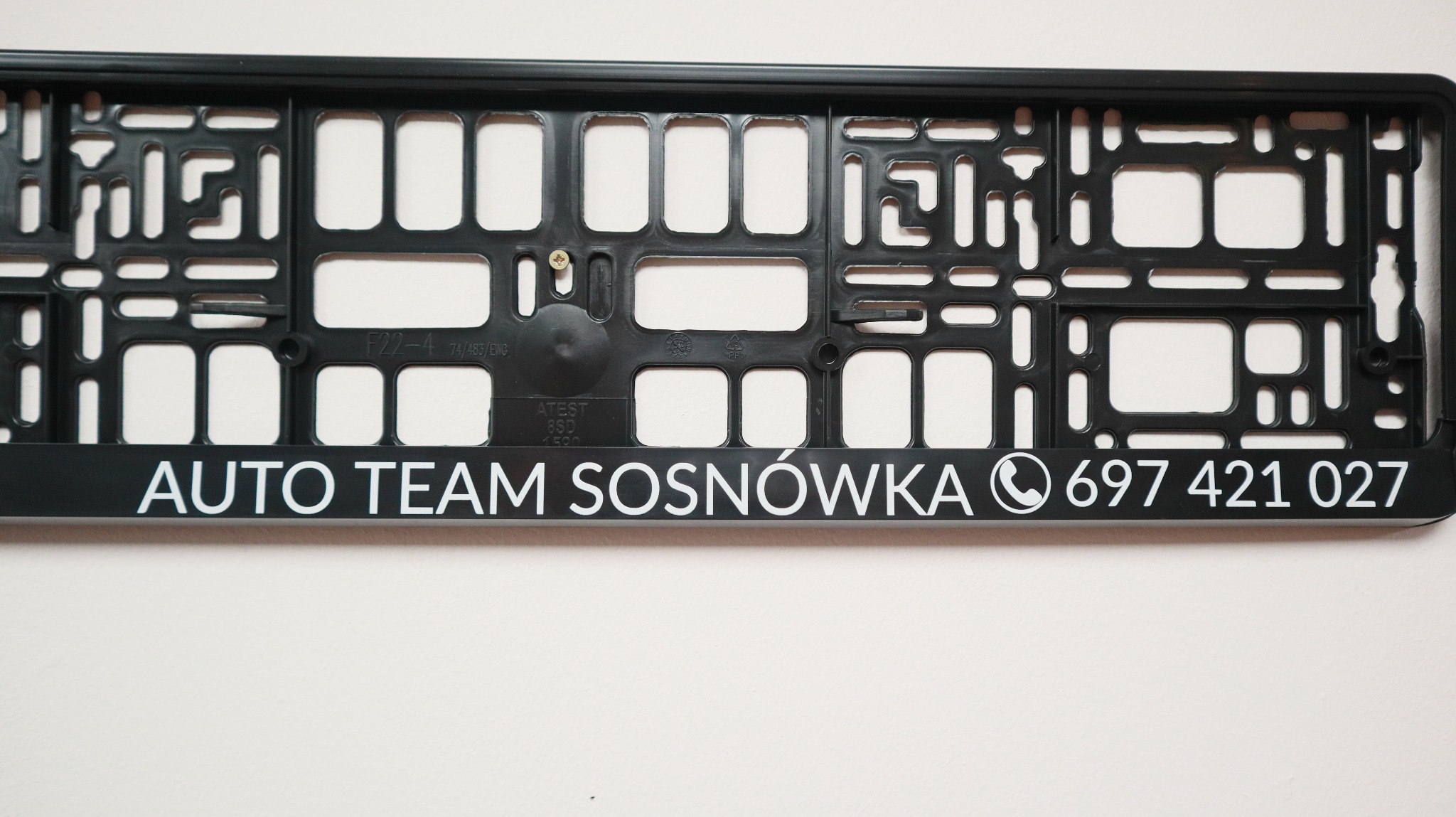 Czarna ramka pod tablicę rejestracyjną z białym nadrukiem 'AUTO TEAM SOSNÓWKA' i numerem telefonu, widoczna z bliska na jasnym tle.