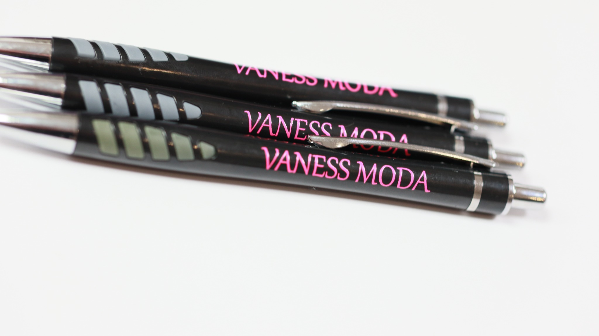 Cztery czarne długopisy z różowym nadrukiem 'Vaness Moda', ułożone równolegle na białym tle, widoczne detale srebrnych elementów i tekstu.