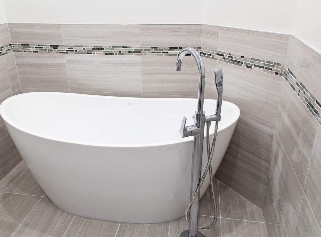 Wolnostojąca wanna z chromowaną baterią podłogową i mozaiką na ścianie z płytek imitujących drewno.