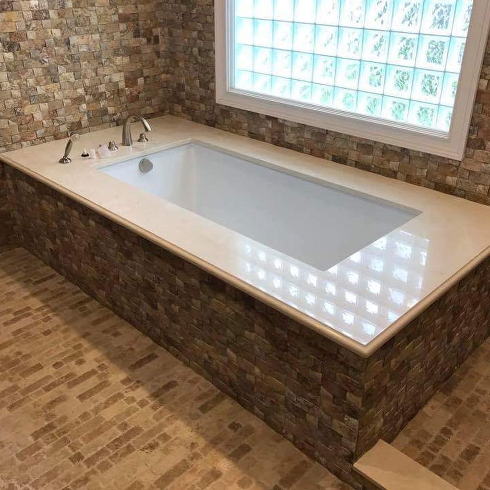 Wanna w obudowie z mozaiki kamiennej, z blatem z jasnego kamienia, bateria umywalkowa w stylu retro, okno z luksferami, podłoga wyłożona płytkami imitującymi kamień.