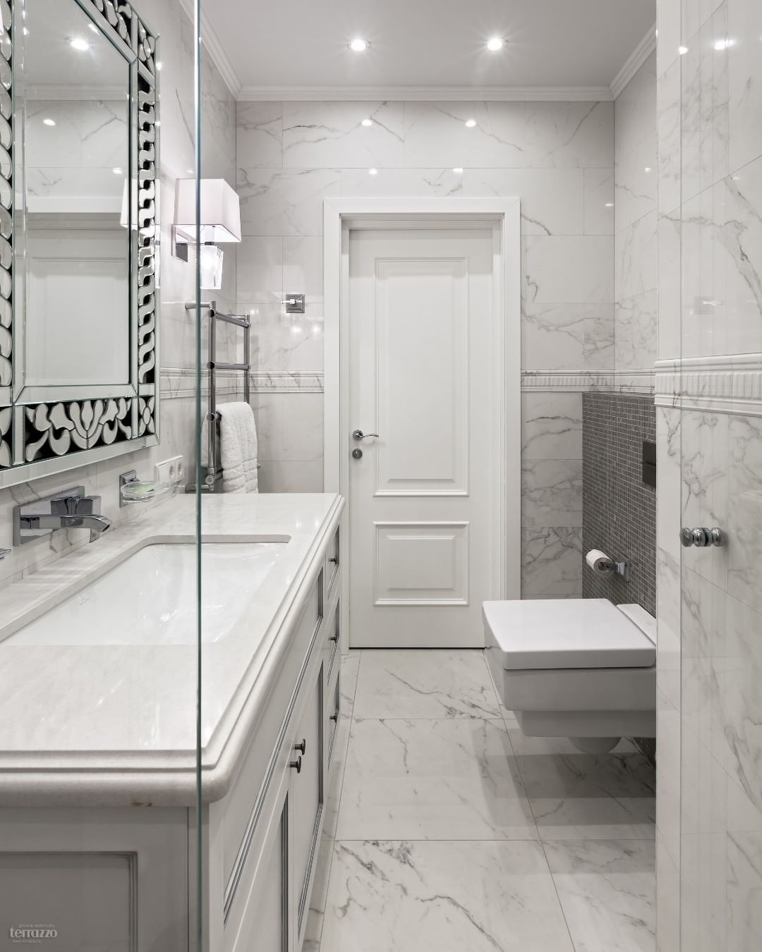 Nowoczesna łazienka z marmurowymi płytkami, dużym lustrem w ozdobnej ramie, wiszącą toaletą i szklaną kabiną prysznicową.