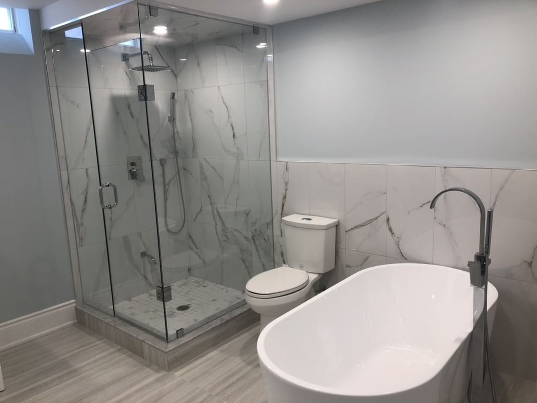 Nowoczesna łazienka z kabiną prysznicową ze szklanymi drzwiami, marmurowymi płytkami, wolnostojącą białą wanną i toaletą.