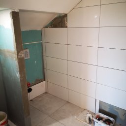 Usługi remontowo budowlane - Wnętrze łazienki w trakcie prac wykończeniowych: częściowo ułożone białe płytki na ścianach pod skosem, widoczna płyta karton-gips na fragmencie ściany, wiadro i pojemnik z narzędziami na podłodze.
