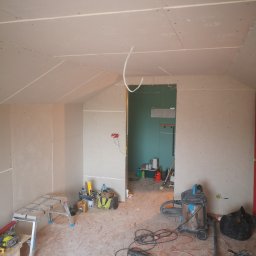 Usługi remontowo budowlane - Poddasze w trakcie zabudowy płytami gipsowo-kartonowymi, widoczne narzędzia, drabina, odkurzacz przemysłowy i przewody elektryczne, ściany i sufit częściowo wykończone, otwór drzwiowy prowadzący...