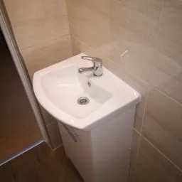 Nowoczesna, narożna umywalka z szafką w łazience wykończonej płytkami imitującymi drewno, chromowana bateria i odpływ.