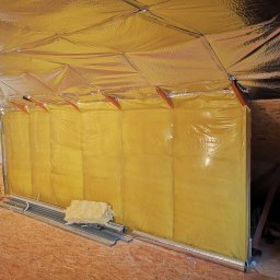 Usługi remontowo budowlane - Izolacja termiczna poddasza folią aluminiową i wełną mineralną, widoczne profile montażowe na drewnianej podłodze.