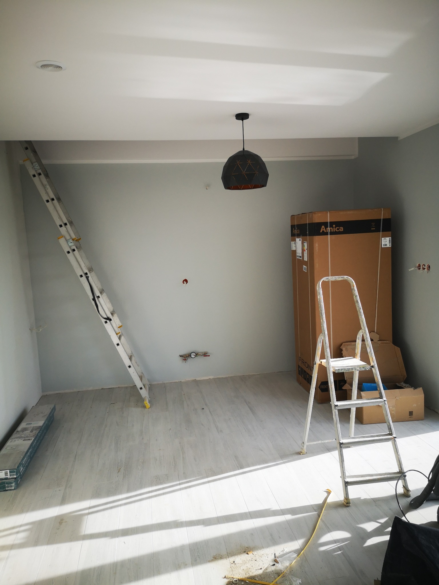 Pokój w trakcie prac wykończeniowych: drabina oparta o ścianę, kartonowe opakowanie sprzętu AGD, zamontowana lampa sufitowa, panele podłogowe czekające na montaż, widoczne ślady po instalacjach...