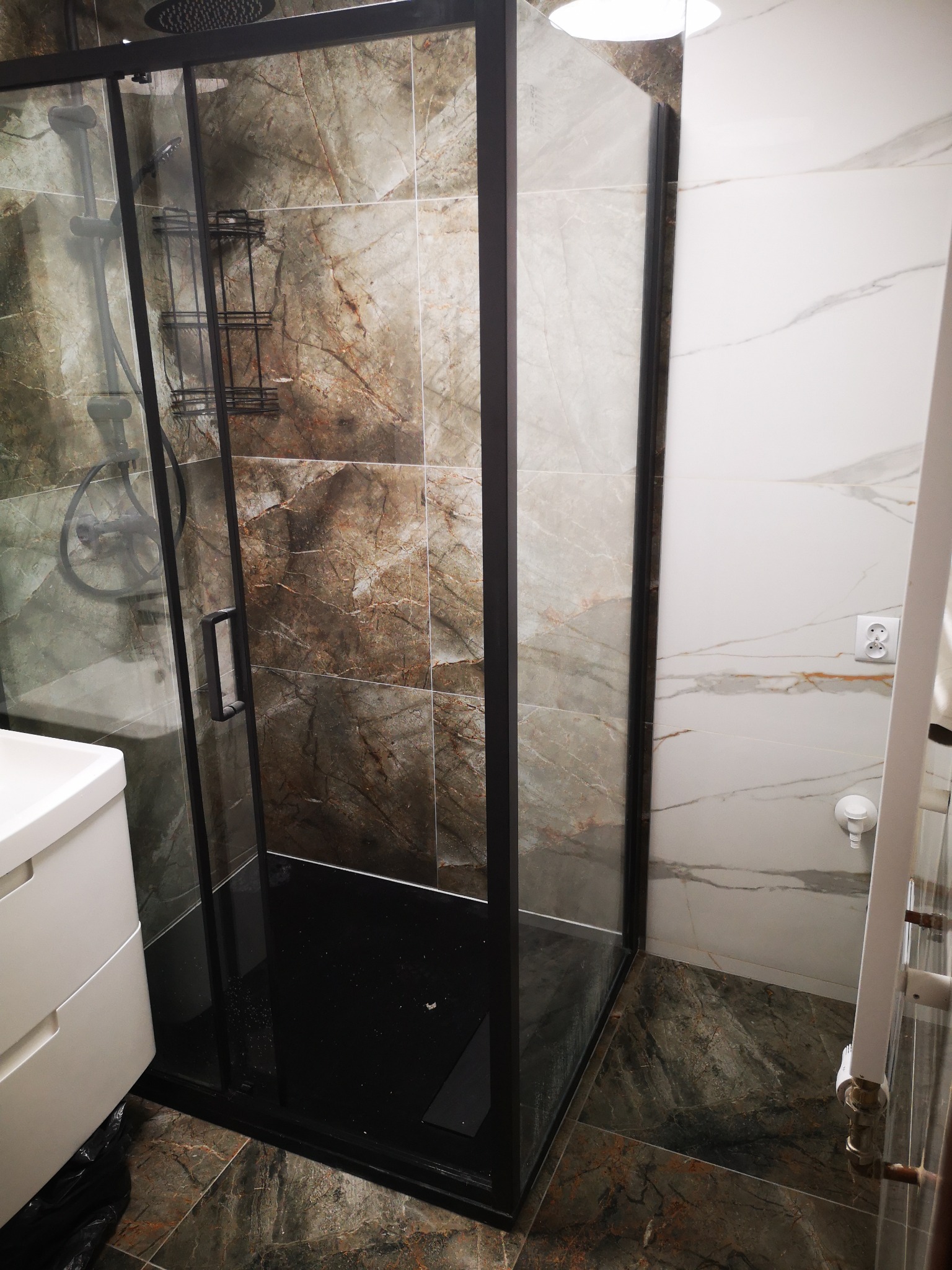 Kabina prysznicowa z czarną ramą i deszczownicą, obłożona płytkami imitującymi marmur w odcieniach brązu i szarości, z czarnym brodzikiem.