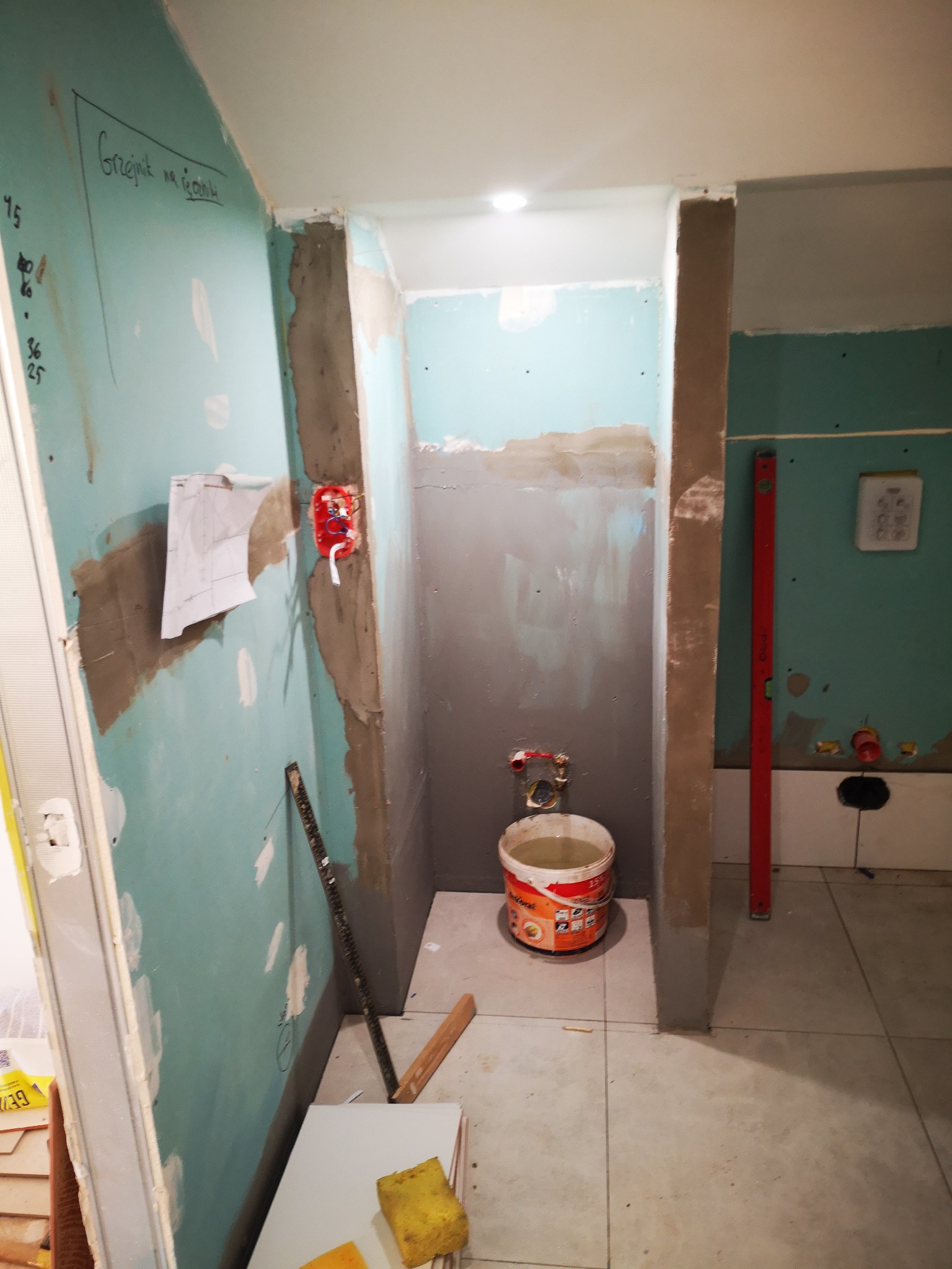 Wnętrze łazienki w trakcie prac przygotowawczych do położenia płytek, widoczne ściany pokryte płytami gipsowo-kartonowymi z zaznaczonymi punktami instalacji, wiadro z zaprawą oraz poziomica oparta...
