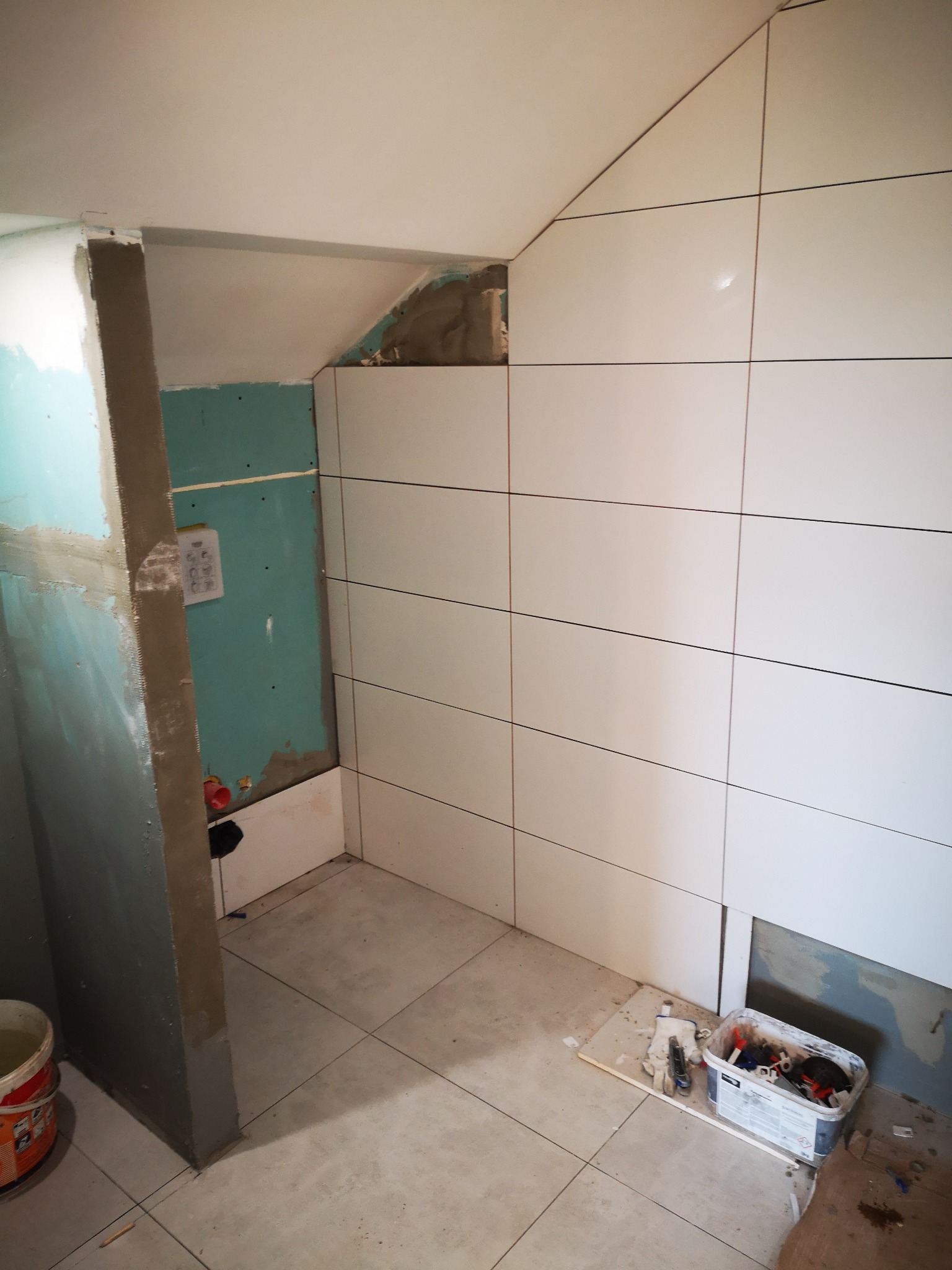 Wnętrze łazienki w trakcie prac wykończeniowych: częściowo ułożone białe płytki na ścianach pod skosem, widoczna płyta karton-gips na fragmencie ściany, wiadro i pojemnik z narzędziami na podłodze.