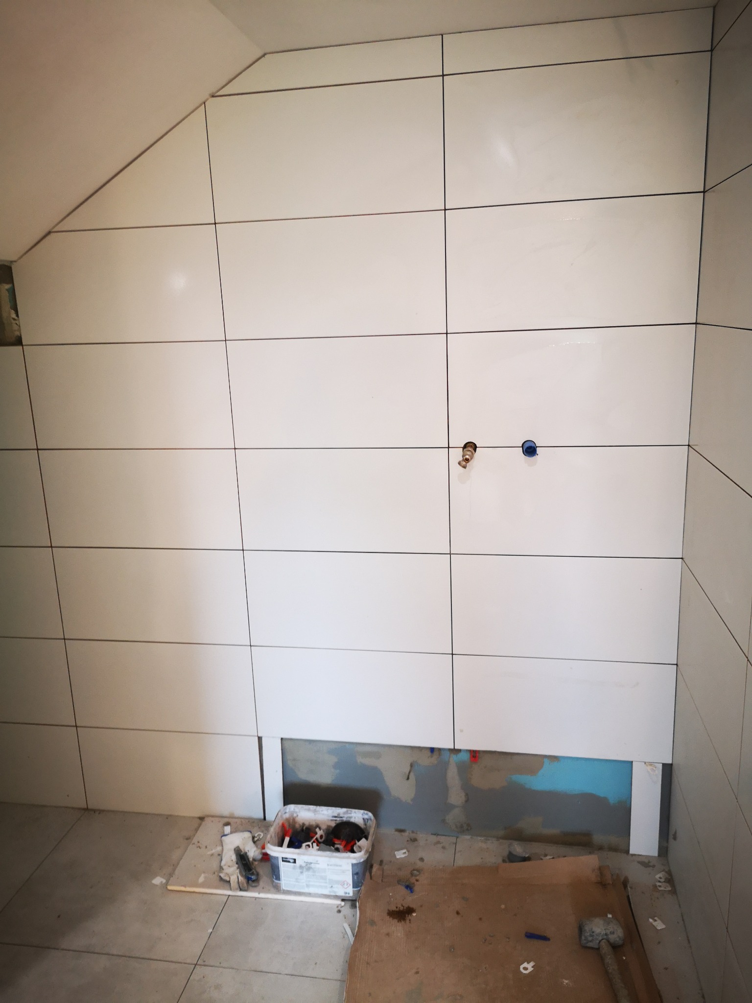 Układanie białych, prostokątnych płytek na ścianie ze skosem, widoczne otwory na instalacje, częściowo odkryta ściana pod płytkami, narzędzia i pojemnik z akcesoriami na podłodze.