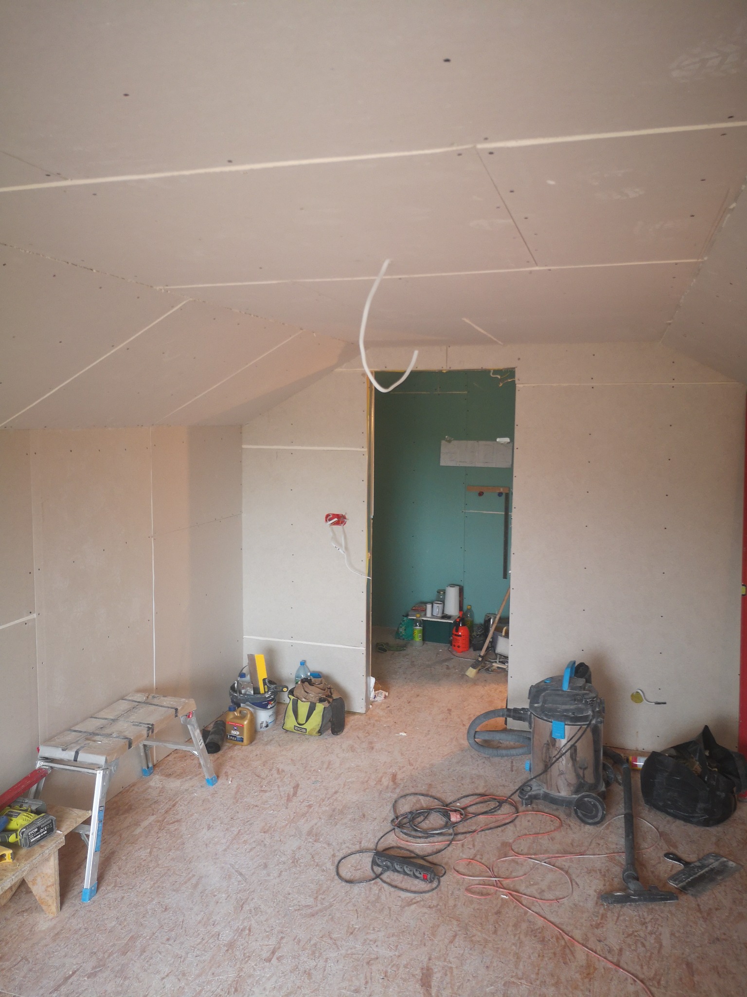 Poddasze w trakcie zabudowy płytami gipsowo-kartonowymi, widoczne narzędzia, drabina, odkurzacz przemysłowy i przewody elektryczne, ściany i sufit częściowo wykończone, otwór drzwiowy prowadzący...