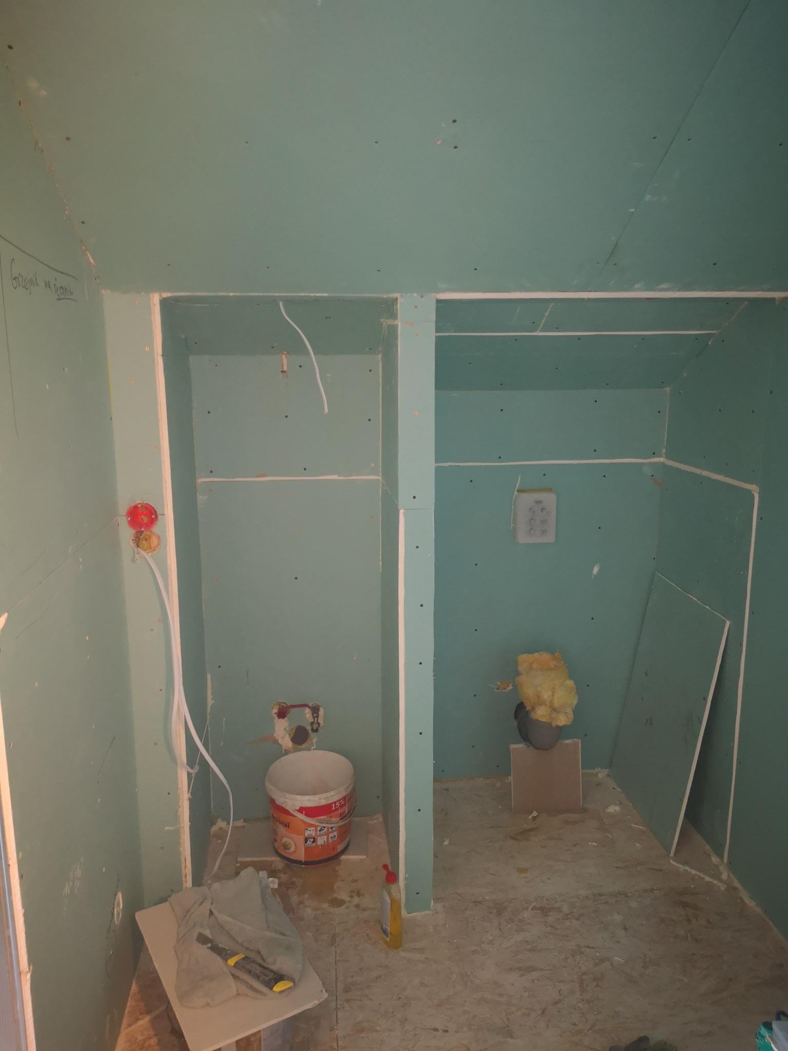 Dwa surowe, oddzielone przestrzenie w pomieszczeniu wyłożonym płytami gipsowo-kartonowymi, widoczne instalacje elektryczne, wiadro z zaprawą, narzędzia i izolacja, etap prac wykończeniowych.