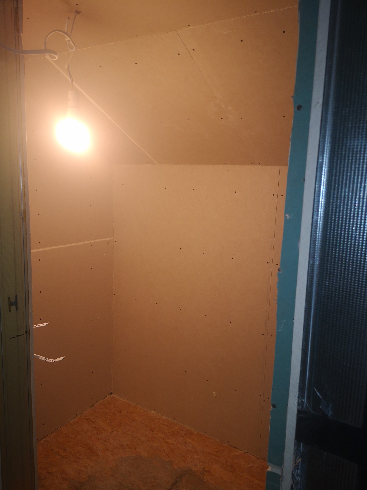 Wnętrze małego pomieszczenia w trakcie prac wykończeniowych, ściany wyłożone płytami kartonowo-gipsowymi, widoczne ślady po wkrętach, podłoga z płyt OSB, oświetlenie stanowi wisząca żarówka.