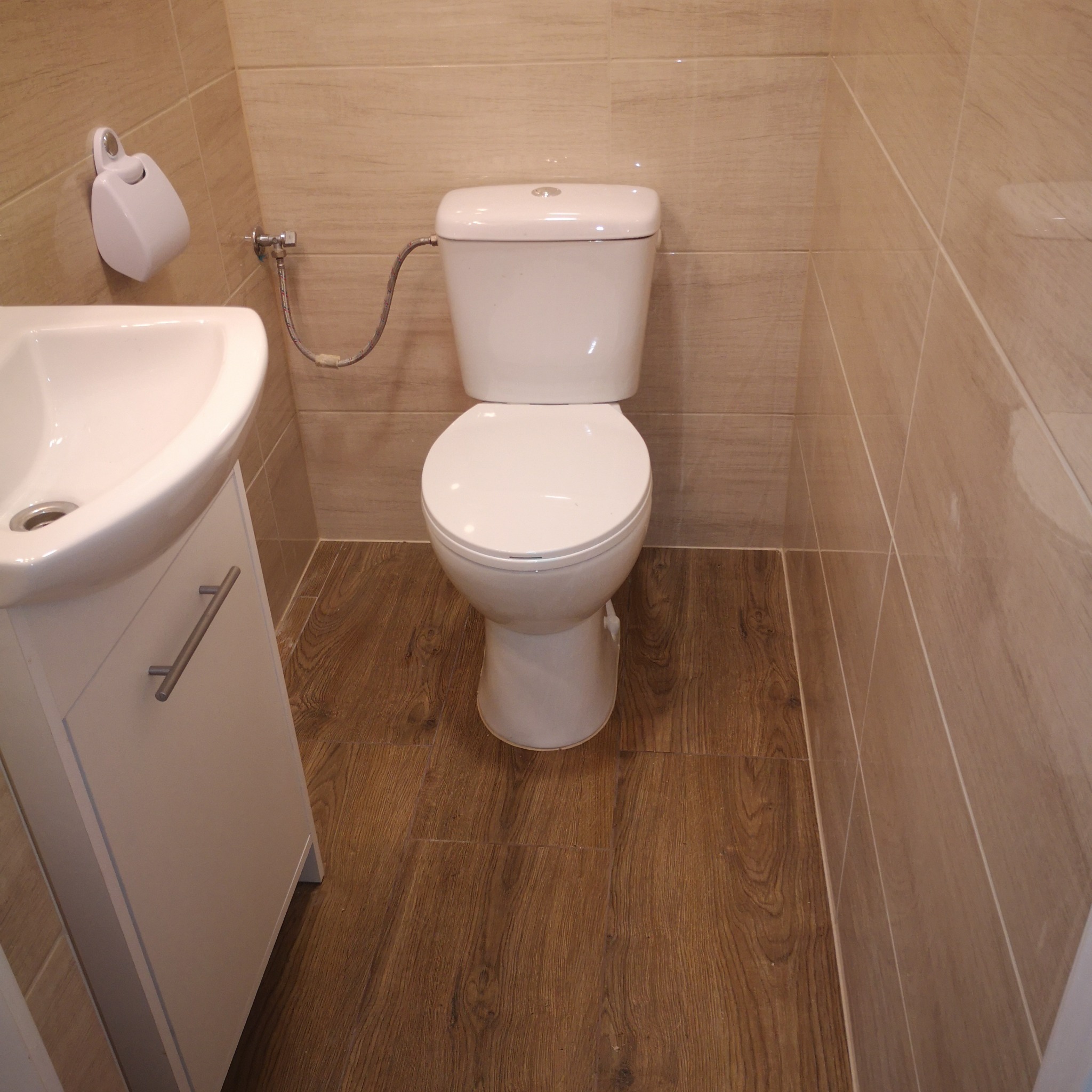 Wykończona łazienka z beżowymi płytkami imitującymi drewno na ścianach i podłodze, biała toaleta i umywalka z szafką.