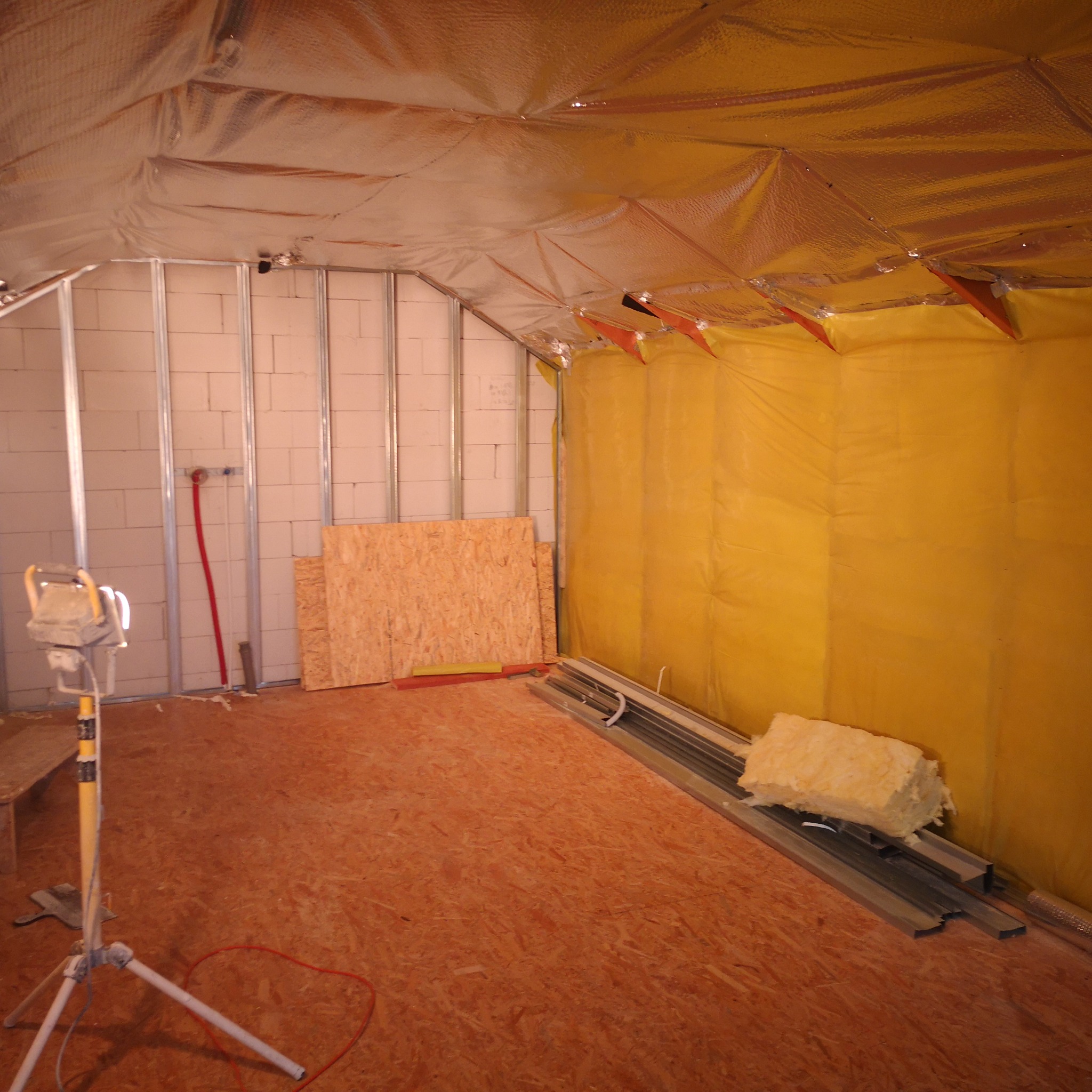 Poddasze w trakcie adaptacji: widoczne ocieplenie folią aluminiową, ściany z bloczków, metalowe profile, płyty OSB i oświetlenie robocze na statywie.