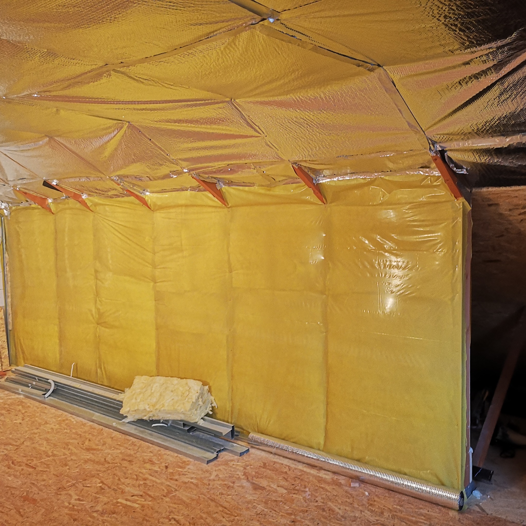 Izolacja termiczna poddasza folią aluminiową i wełną mineralną, widoczne profile montażowe na drewnianej podłodze.