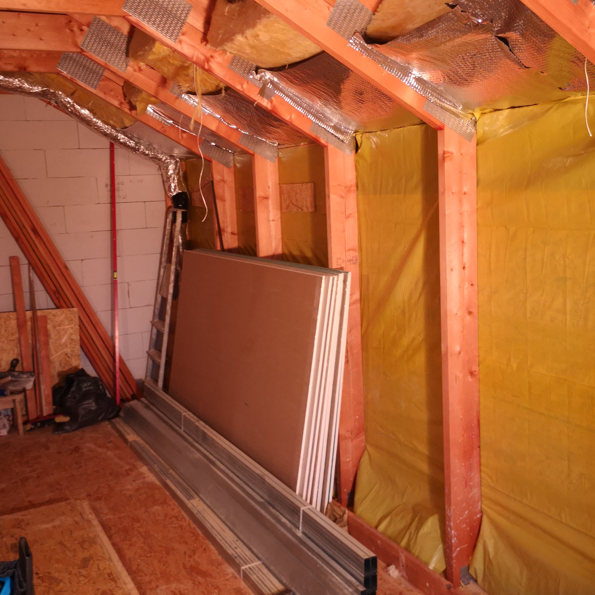 Poddasze w trakcie adaptacji: widoczne ocieplenie folią, drewniana konstrukcja dachu wzmocniona metalowymi łącznikami, płyty gipsowo-kartonowe i profile stalowe przygotowane do montażu na podłodze...