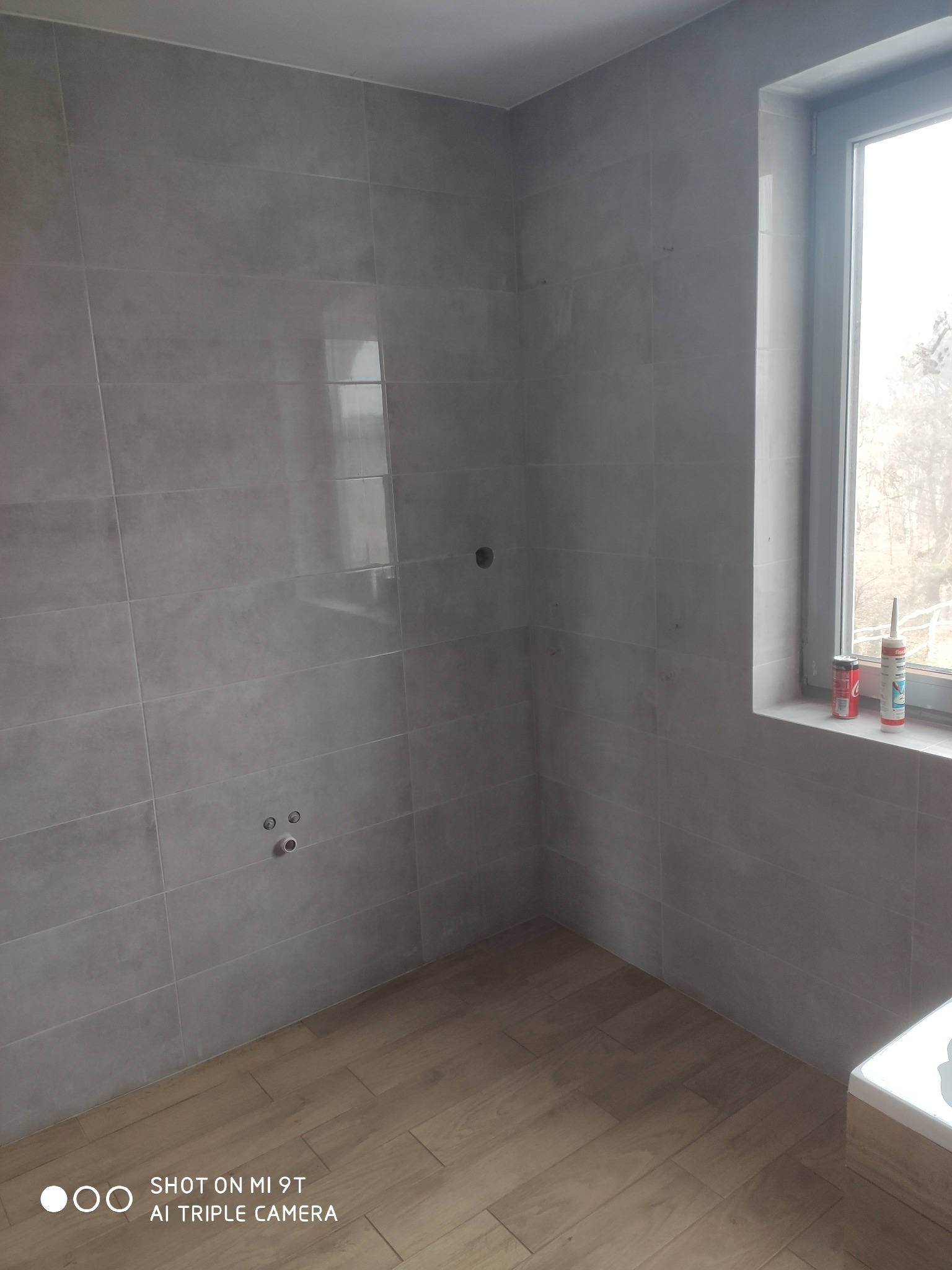 Wnętrze łazienki w trakcie prac wykończeniowych, szare płytki na ścianach, drewnopodobne płytki na podłodze, widoczne otwory na armaturę i okno z silikonami na parapecie.