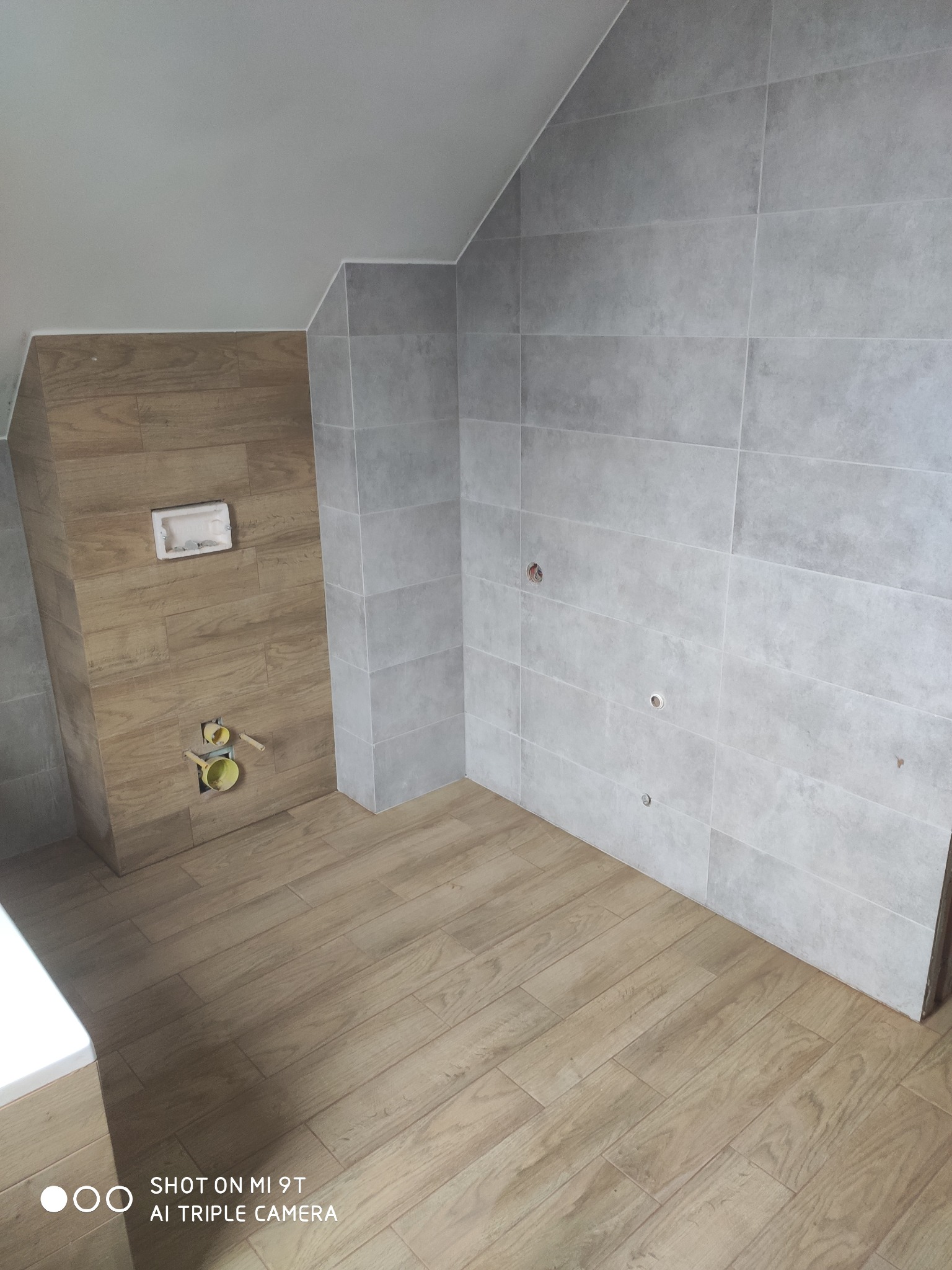 Łazienka w trakcie wykańczania: szare płytki na ścianach, drewnopodobne płytki na podłodze i ścianie, widoczne instalacje hydrauliczne.