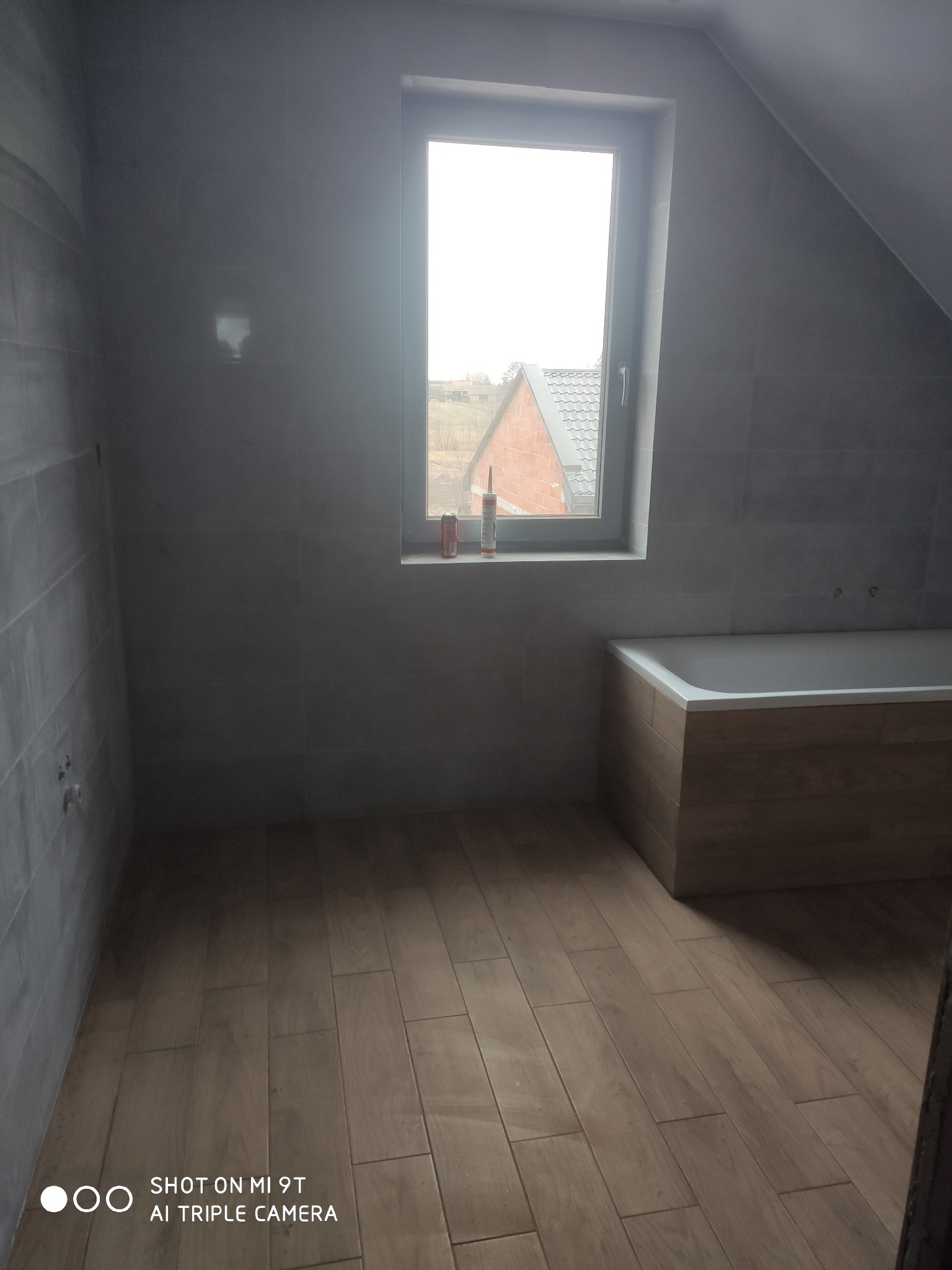 Szara łazienka na poddaszu z drewnopodobnymi płytkami na podłodze i obudowie wanny, widoczna tuba silikonu na parapecie okna.