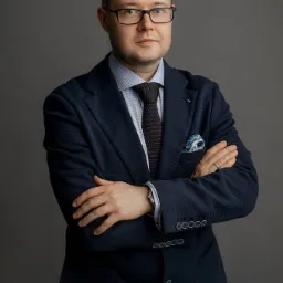 Portret mężczyzny w okularach, ciemnym garniturze i wzorzystym krawacie, stojącego na szarym tle z założonymi rękami.