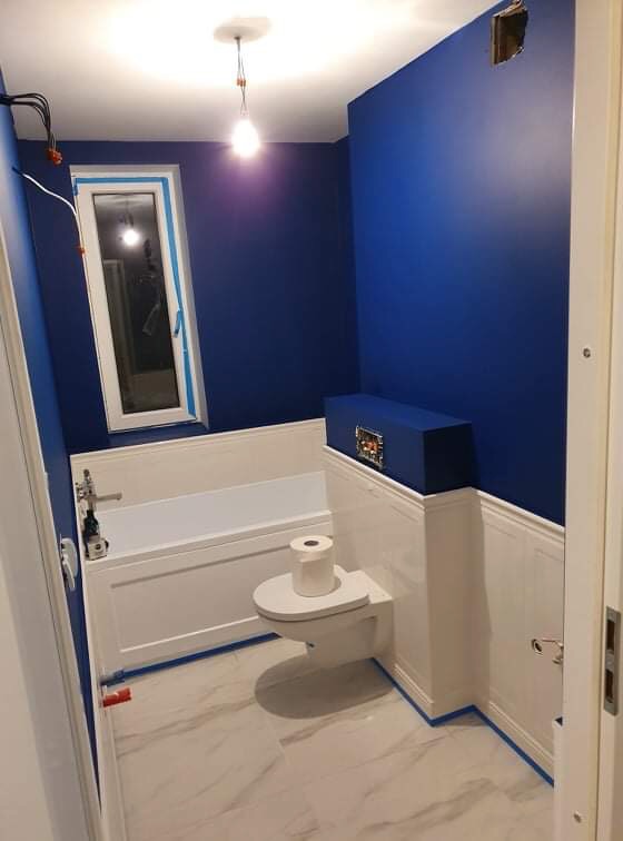 Niebieska łazienka w trakcie wykańczania: pomalowane ściany, biała wanna, podwieszana toaleta z rolką papieru, marmurowe płytki podłogowe, widoczne taśmy malarskie i niedokończone elementy...