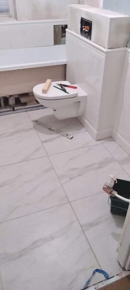 Świeżo wykafelkowana podłoga w łazience z zamontowaną podwieszaną toaletą, na której leżą narzędzia i kawałek drewna, widoczne elementy instalacji w ścianie oraz przygotowana wanna do obudowy.