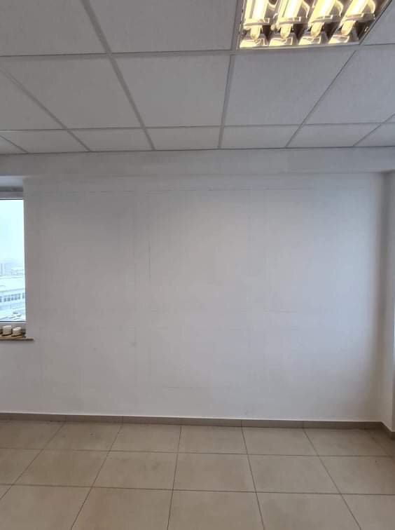 Pusta, pomalowana na biało ściana w pomieszczeniu biurowym z widocznym oknem i kafelkami na podłodze, przygotowana do dalszych prac wykończeniowych.