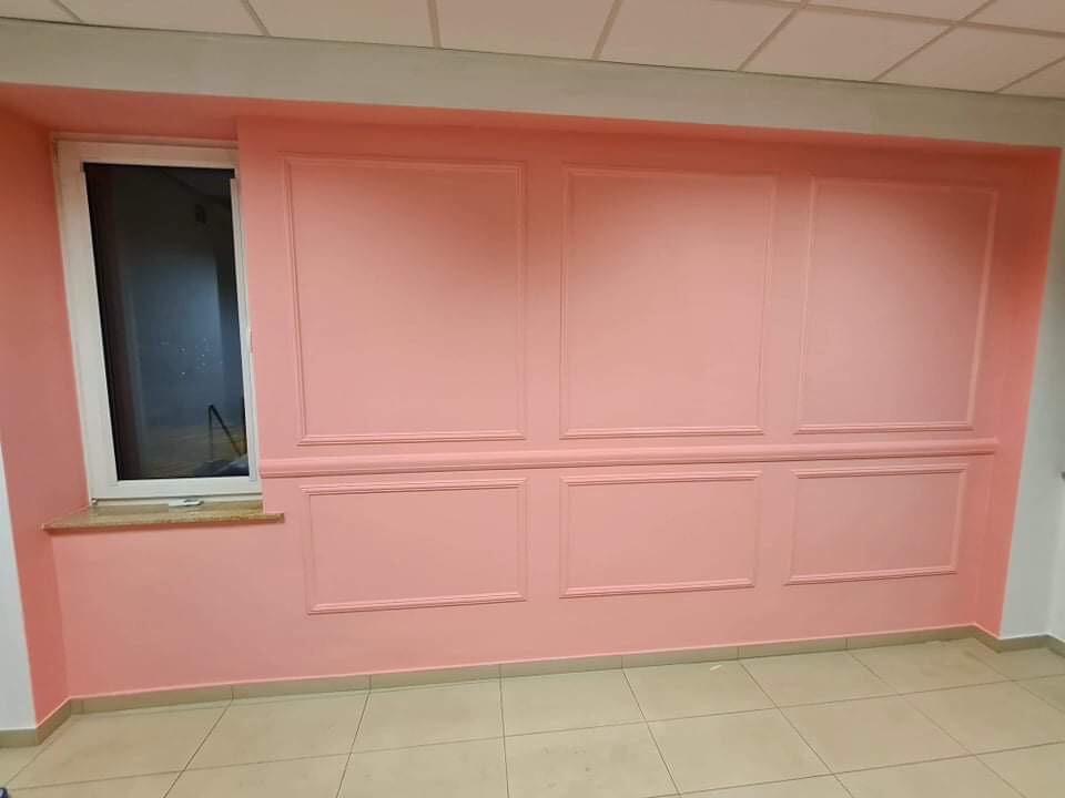 Ściana w kolorze pudrowego różu z dekoracyjnymi panelami i listwami, widok na okno z marmurowym parapetem, kremowa podłoga z płytek.