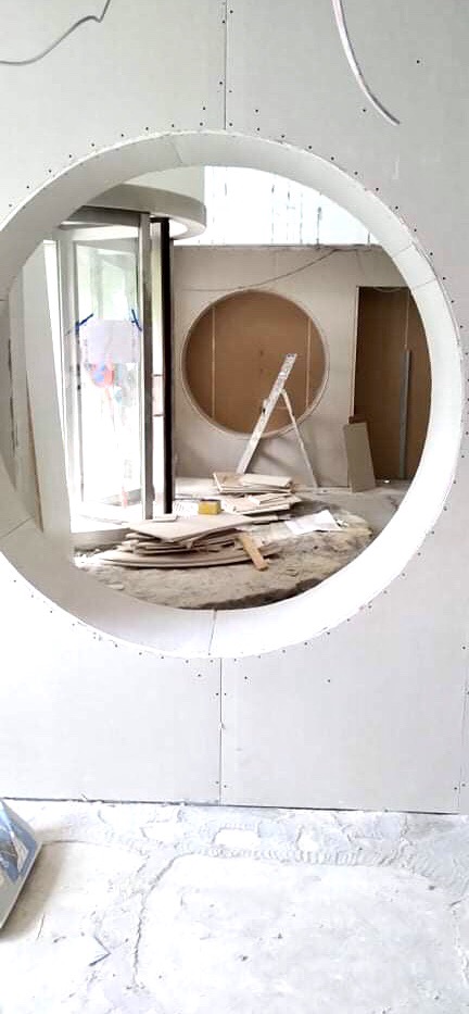 Wnętrze w trakcie prac wykończeniowych z okrągłym otworem w ścianie z płyt gipsowo-kartonowych, widoczna drabina oparta o ścianę z kolejnym okrągłym otworem, porozrzucane płyty i narzędzia.