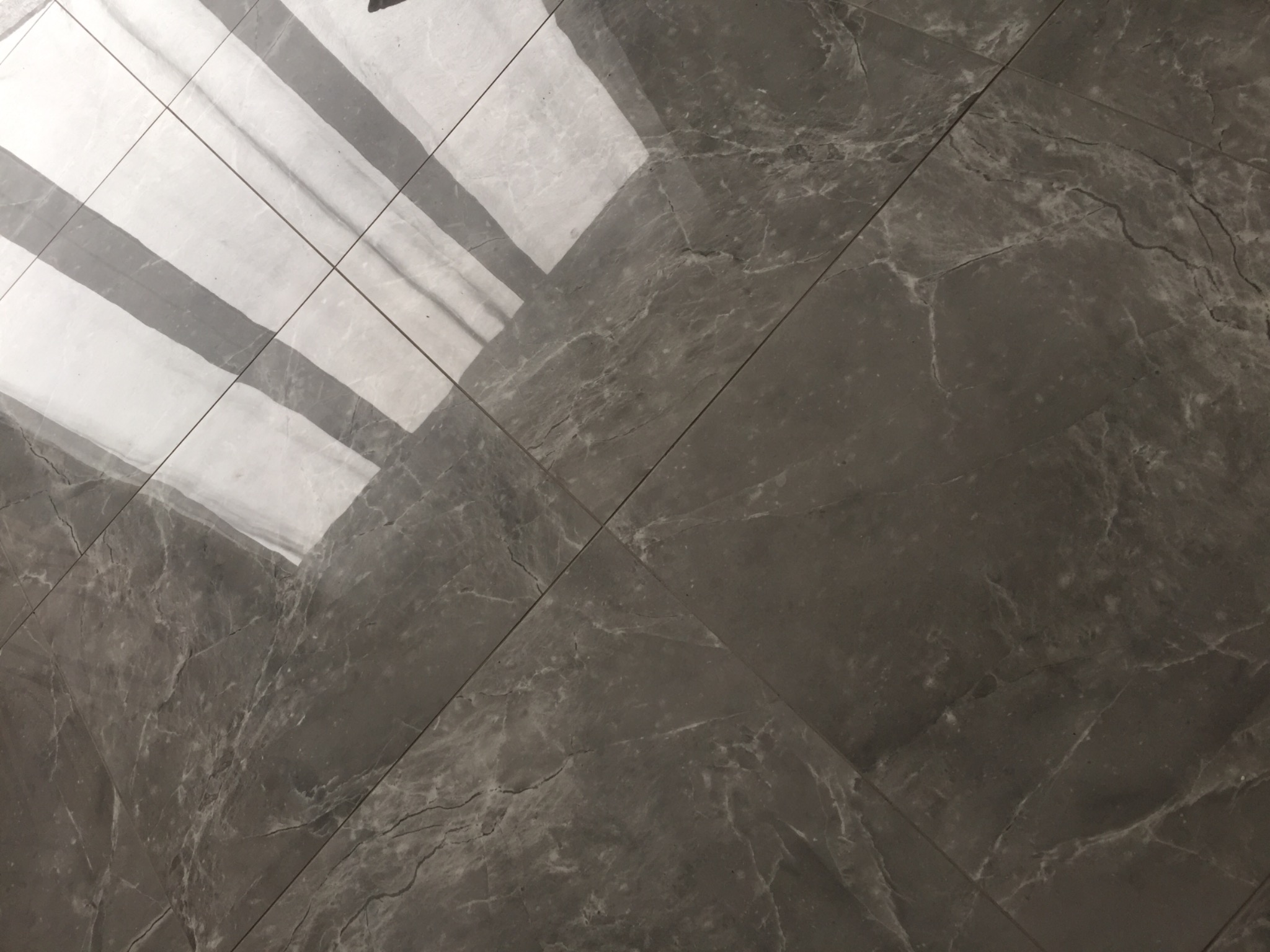 Szare, błyszczące płytki podłogowe imitujące marmur, ułożone pod kątem, z widocznym odbiciem okna.
