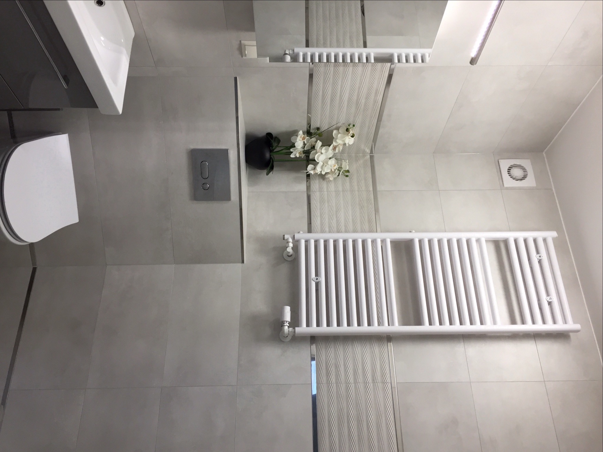 Nowoczesna łazienka wykończona w szarościach z białym grzejnikiem drabinkowym, wiszącą umywalką i toaletą. Dekoracyjny akcent stanowi czarny wazon z białymi kwiatami. Widoczna wentylacja...