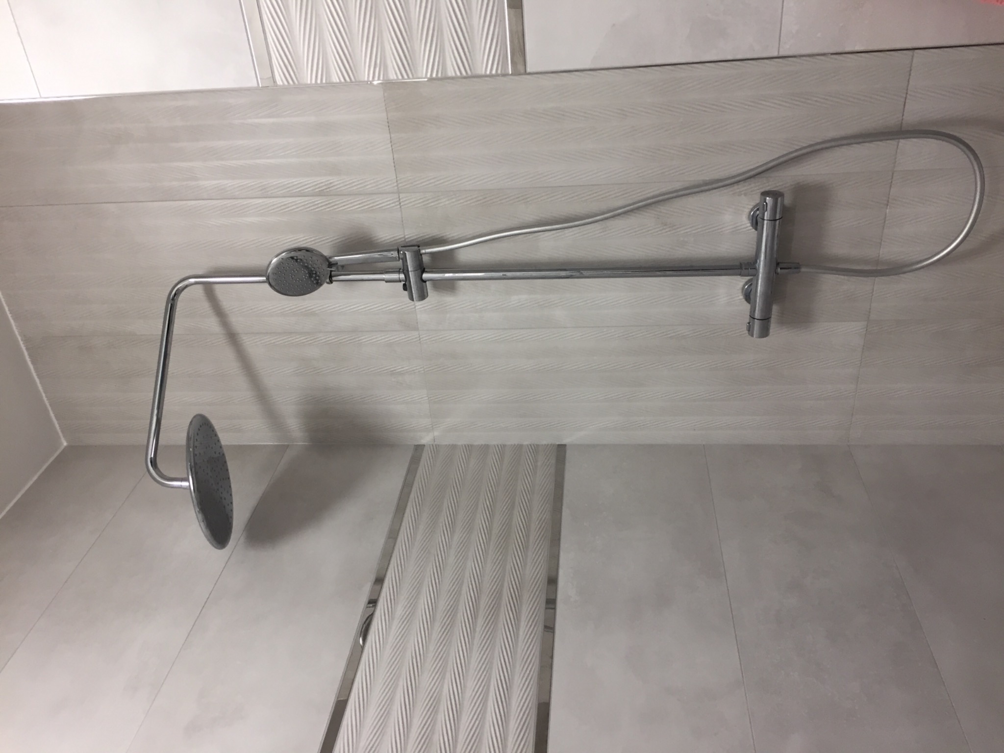 Nowoczesny, chromowany zestaw prysznicowy z deszczownicą i słuchawką, zamontowany na jasnych, wzorzystych płytkach w łazience.