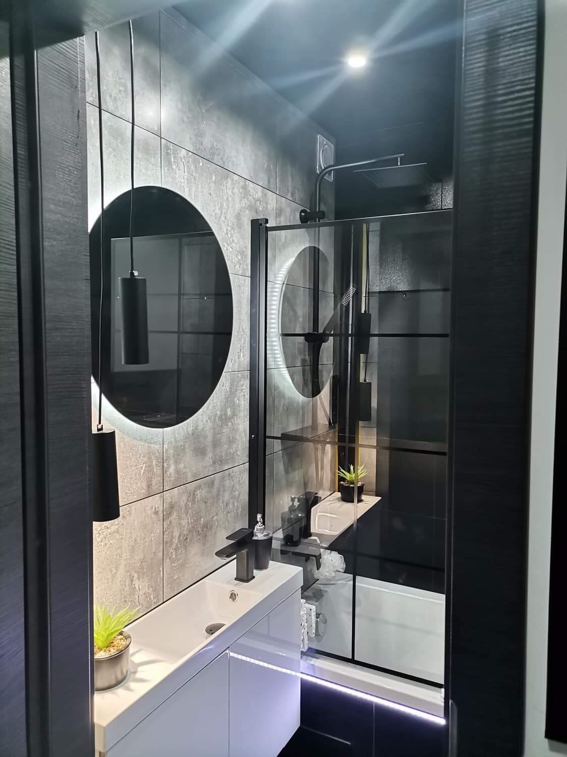 Nowoczesna łazienka z szarymi płytkami imitującymi beton, okrągłym lustrem z podświetleniem LED, czarną armaturą, białą umywalką z szafką i kabiną prysznicową z czarnymi ramami.