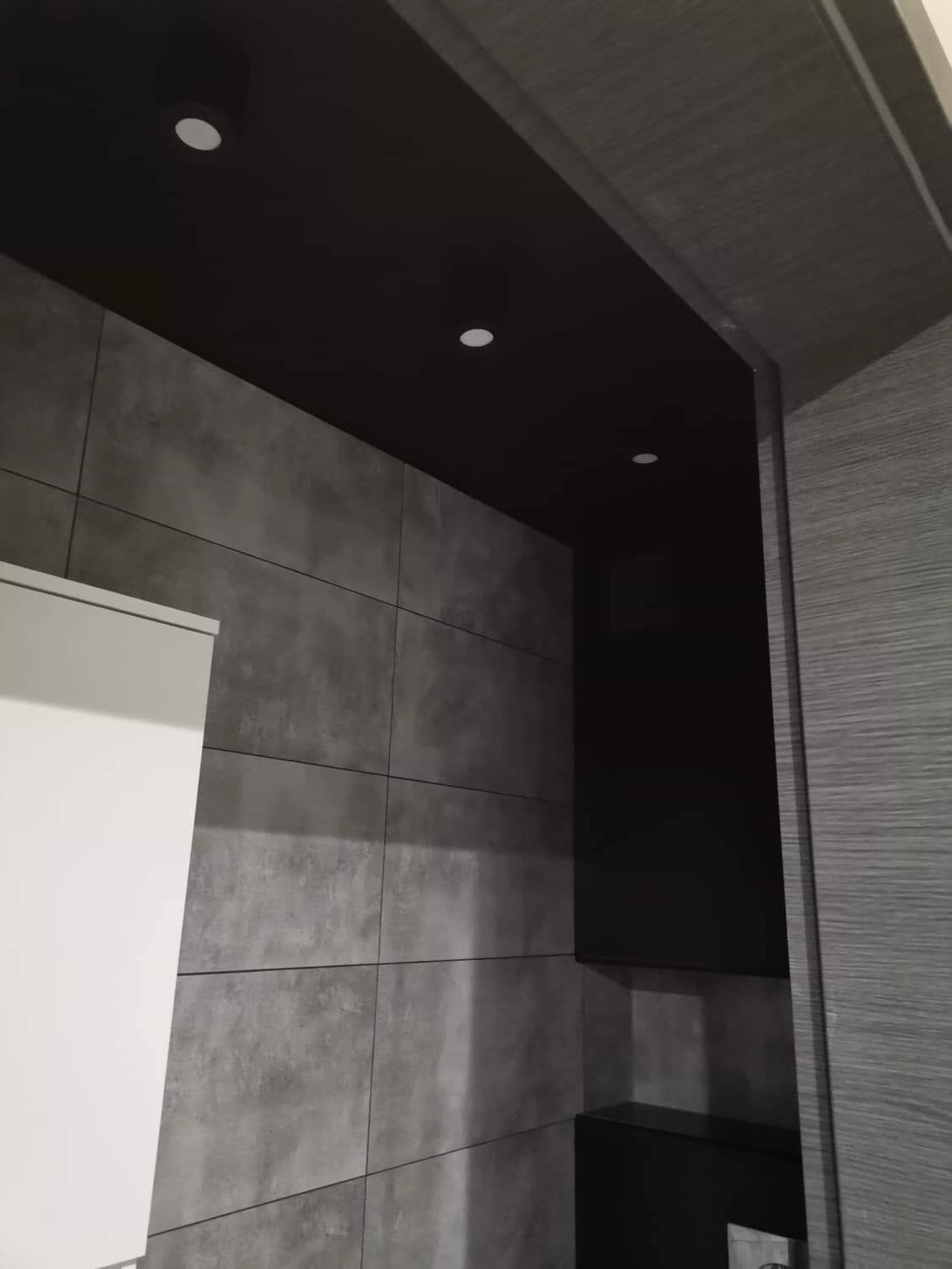 Szara łazienka z ciemnym sufitem i punktowym oświetleniem, widok zza drewnianej framugi drzwi.