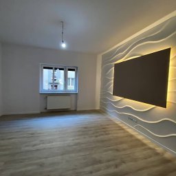 ZAO TEAM SPÓŁKA Z OGRANICZONĄ ODPOWIEDZIALNOŚCIĄ - Pusty pokój po remoncie z panelami 3D na ścianie i podświetleniem LED. Widoczne okno z grzejnikiem i wisząca żarówka. Podłoga wyłożona panelami imitującymi drewno.