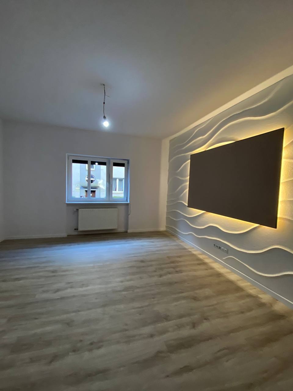 Pusty pokój po remoncie z panelami 3D na ścianie i podświetleniem LED. Widoczne okno z grzejnikiem i wisząca żarówka. Podłoga wyłożona panelami imitującymi drewno.