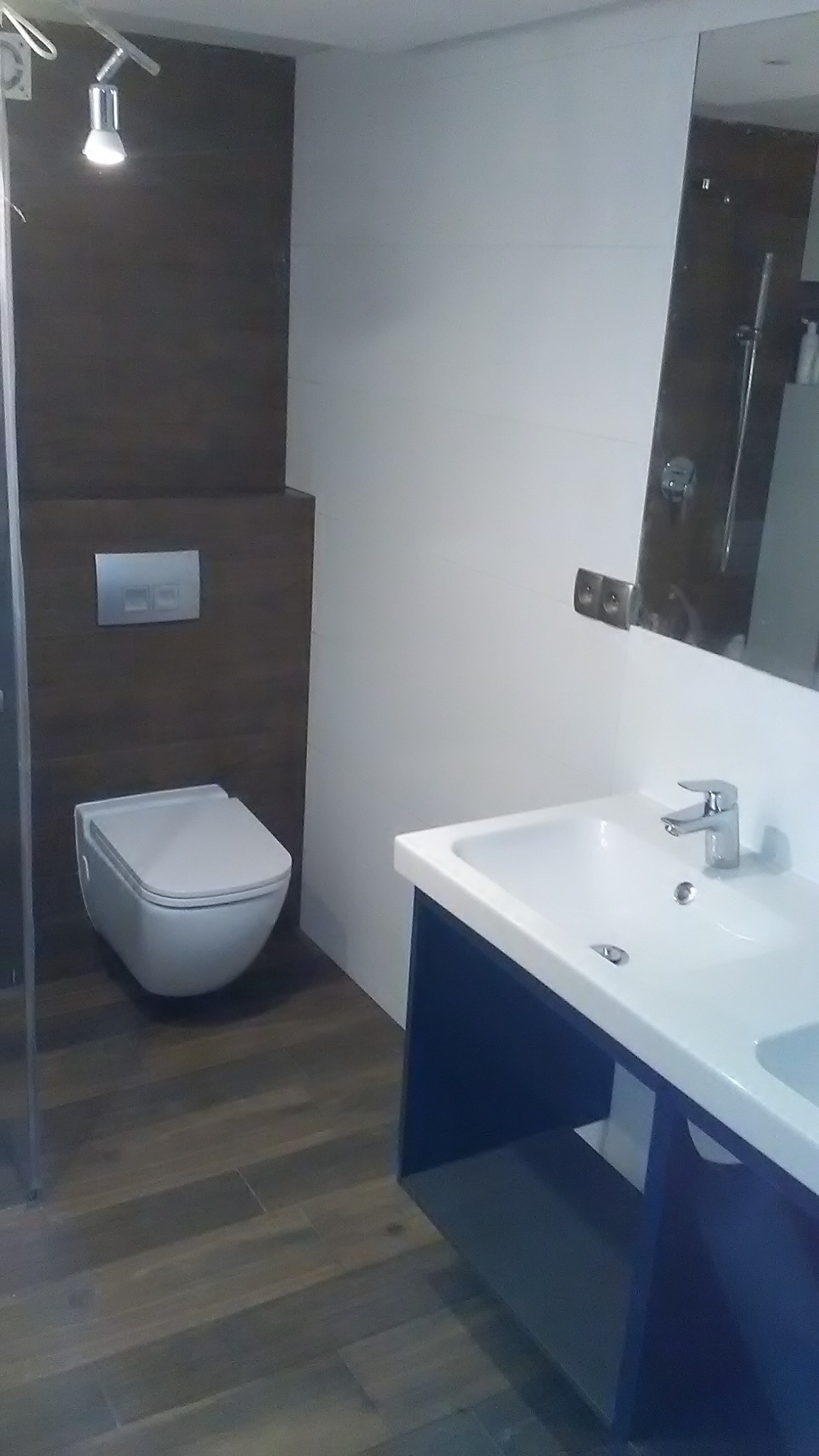 Nowoczesna łazienka po remoncie: podwieszana toaleta, umywalka z szafką w kolorze granatowym, lustro i brązowe płytki podłogowe. Minimalistyczny design.