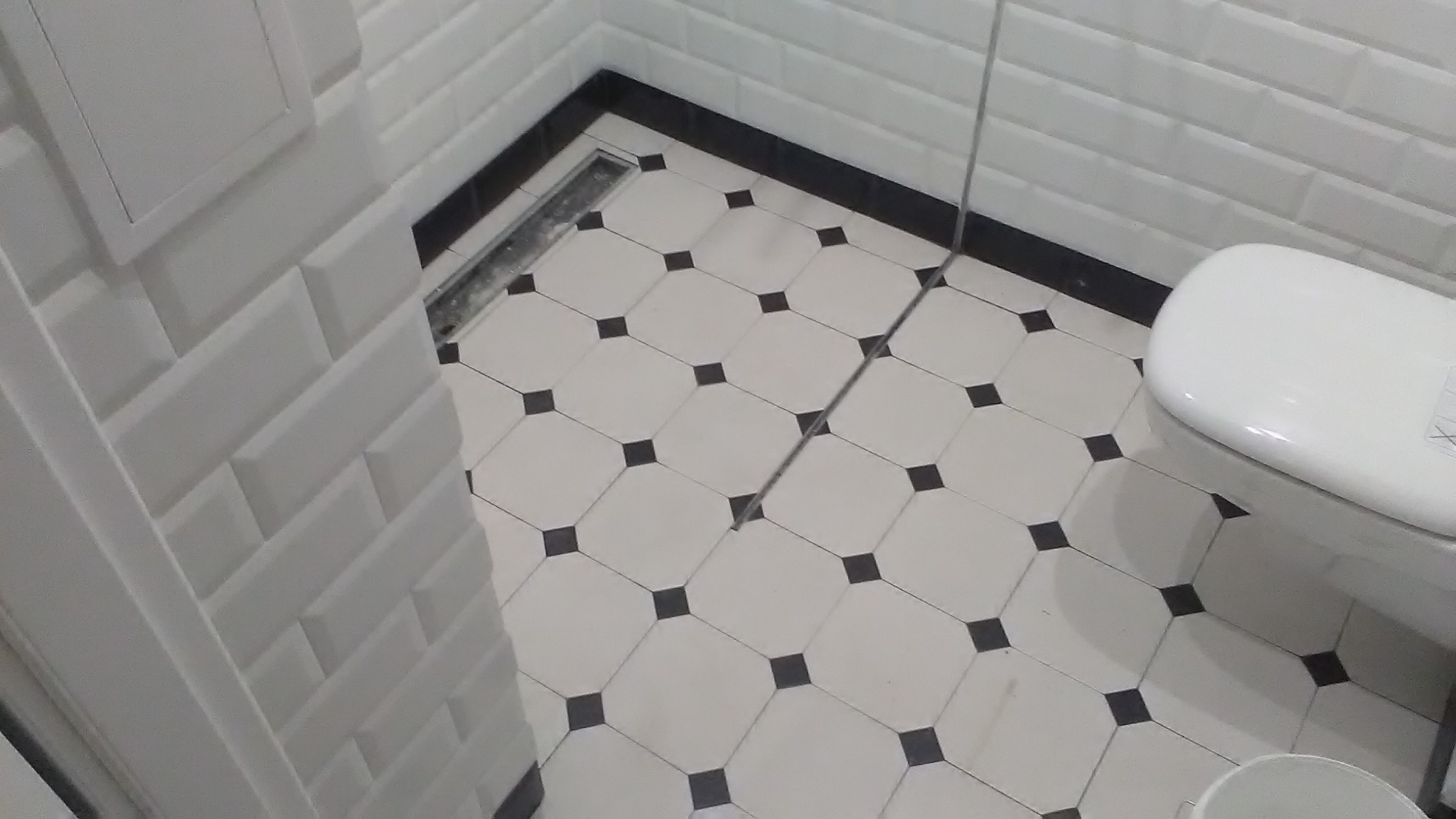 Wykończona łazienka z białą armaturą, czarno-białą podłogą w romby i białymi płytkami typu metro na ścianach. Widoczny odpływ liniowy.