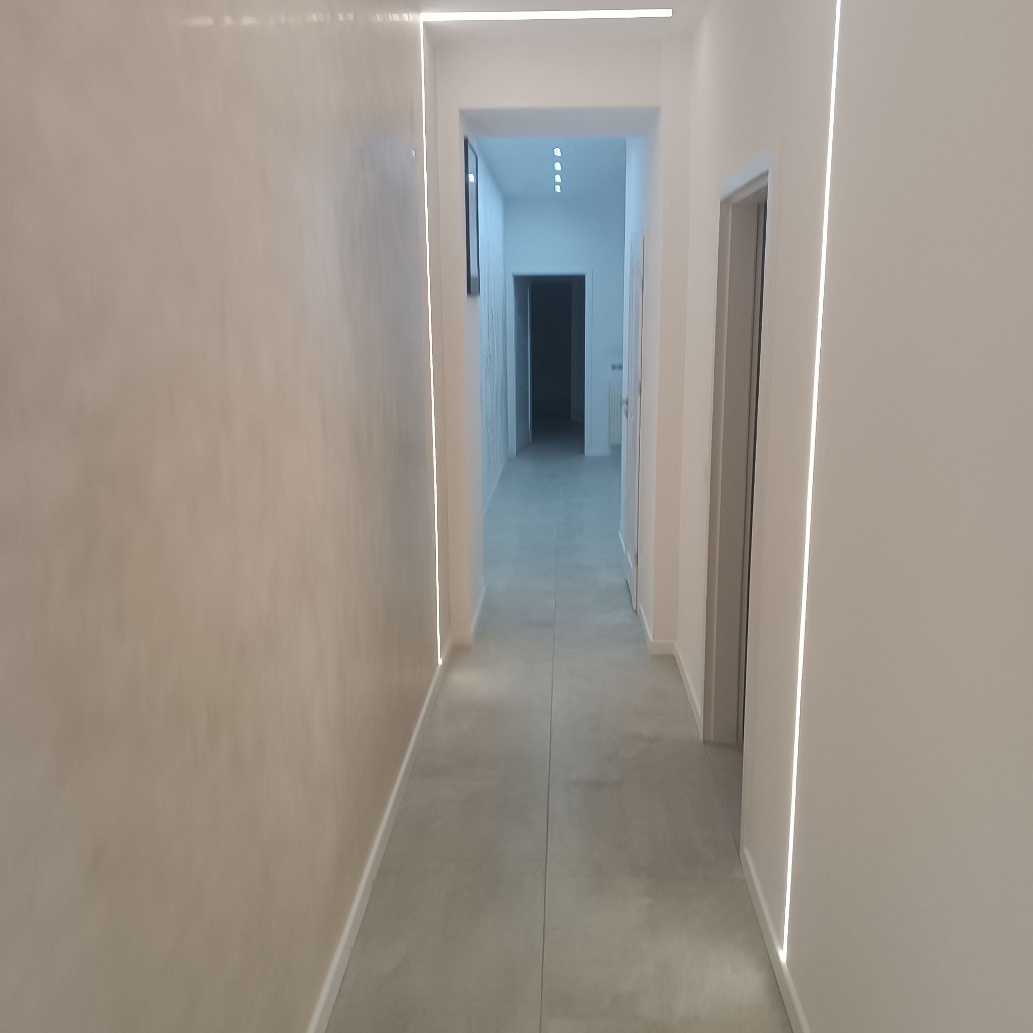 Nowoczesny korytarz z oświetleniem LED wzdłuż ścian i gładzią w odcieniach beżu. Minimalistyczny design z szarą podłogą i widokiem na dalszą część mieszkania.