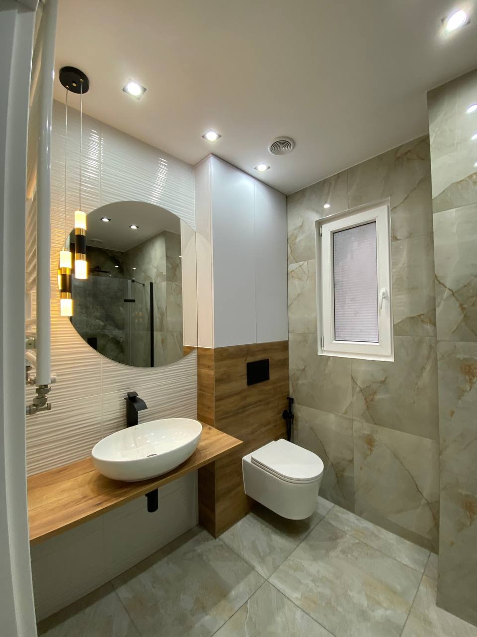 Nowoczesna łazienka z owalną umywalką na drewnianym blacie, okrągłym lustrem i podwieszaną toaletą. Beżowe płytki imitujące marmur na ścianach i podłodze.