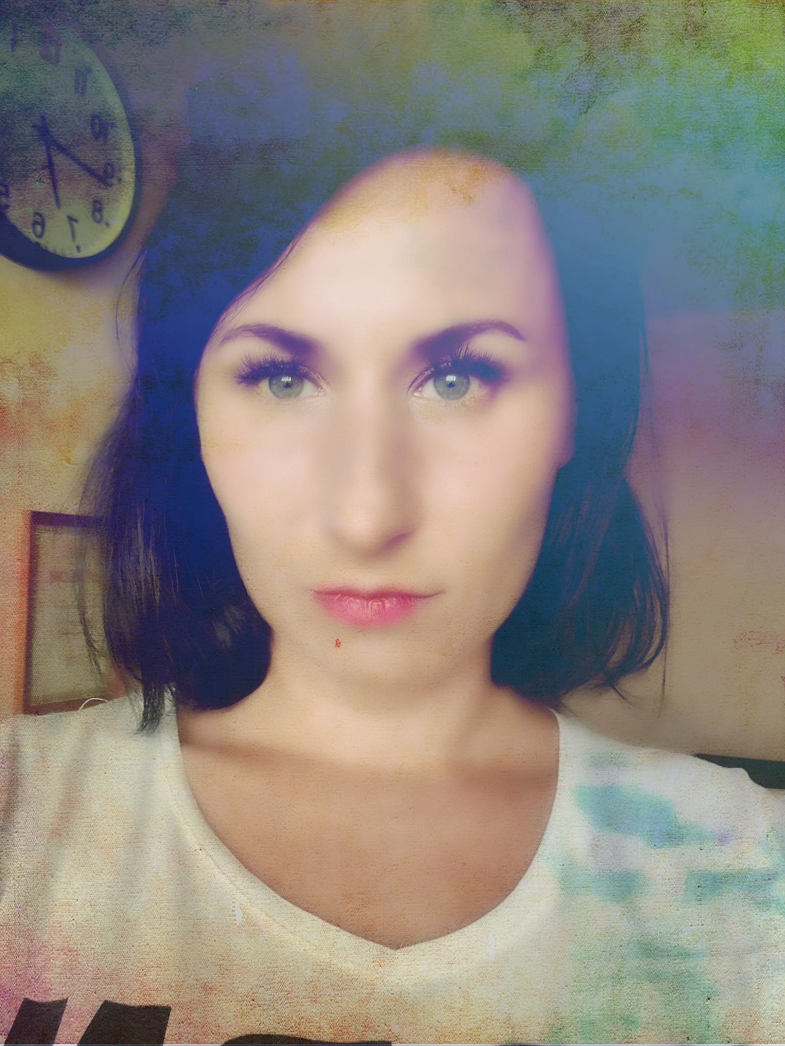 Portret kobiety z krótkimi ciemnymi włosami, zielonymi oczami i delikatnym makijażem, na rozmytym tle widać zegar na ścianie.