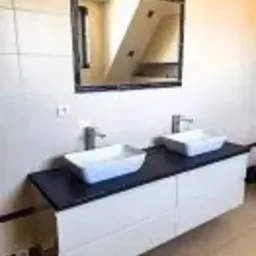 Łazienka z podwójną, prostokątną umywalką nablatową na białej szafce z czarnym blatem, nad którą wisi lustro w ciemnej ramie odbijające fragment schodów.