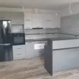 Jasnoszara zabudowa kuchenna z czarną lodówką i piekarnikiem, szary blat, drewnopodobna podłoga, widok na okno, surowy stan wykończenia wnętrza.