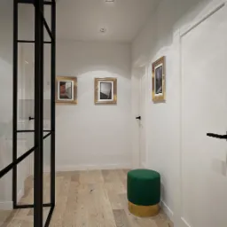 Wnętrze przedpokoju z drewnianą podłogą, białymi ścianami ozdobionymi trzema obrazami w złotych ramach, zielonym pufem na złotym postumencie oraz czarną, przeszkloną ścianką działową z rośliną...