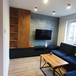 Nowoczesny salon z drewnianą zabudową ściany, czarną szafką RTV, drewnianym stolikiem kawowym na metalowym stelażu i narożnikiem w kolorze granatowym.