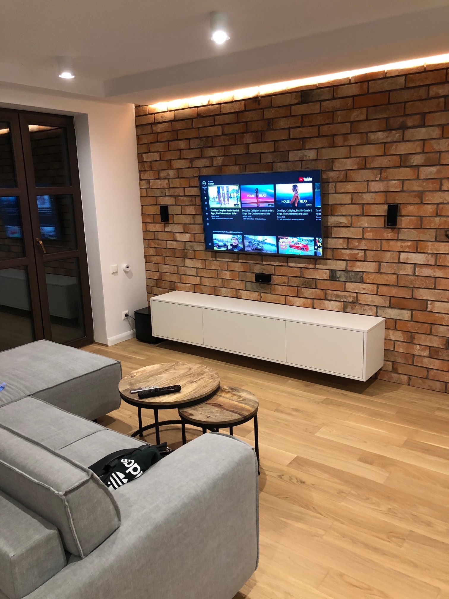 Salon z ceglaną ścianą, na której wisi telewizor, pod nim biała szafka RTV, drewniana podłoga i szara sofa z okrągłymi stolikami kawowymi.
